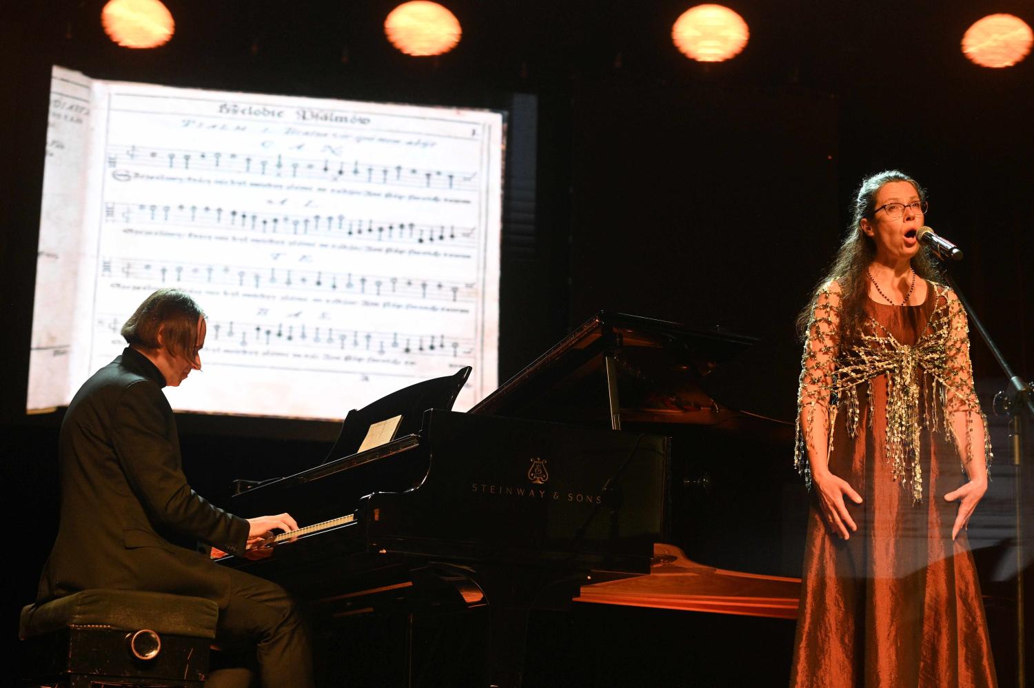 Śpiewaczka Joanna Korpiela-Jatkowska wykonuje utwór z organistą Tomaszem Orlowem grającym na czarnym fortepianie, w tle nuty starego utworu
