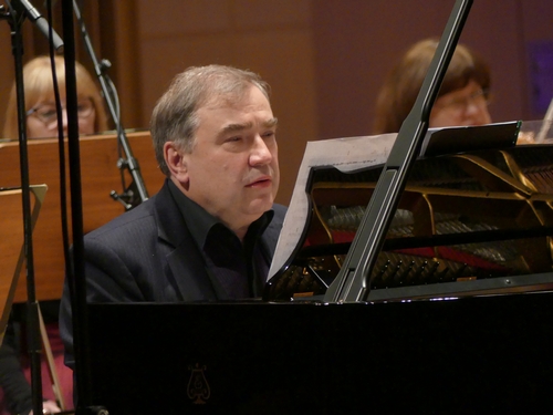 Pianista Marek Mizera siedzi przy czarnym fortepianie, jest ubrany w czarną koszulę, w tle grają muzycy orkiestry
