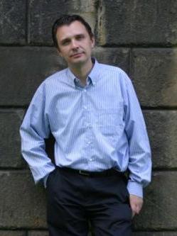 Kompozytor Mikołaj Górecki stoi oparty o ceglany mur, jest ubrany w niebieską koszulę i czarne spodnie