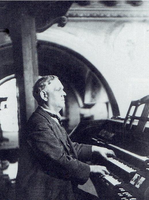 Kompozytor Victor Clariss Czajanek siedzi przy klawiaturze organów, jest ubrany w czarny surdut, ciemny krawat i białą koszulę