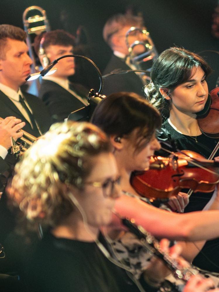 Czterech muzyków z orkiestry gra na skrzypcach, to młodzi, usmiechnięci ludzie, są ubrani w czarne stroje