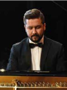 Pianista Jacek Obstarczyk gra na fortepianie, ubrany jest w czarny smoking, białą koszulę oraz czarną muszkę