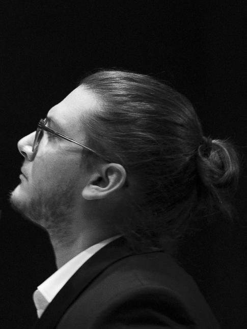 Dyrygent Maciej Cisowski nosi okulary i występuje w czarnym fraku, białej koszuli i z małym kokiem z tyłu głowy