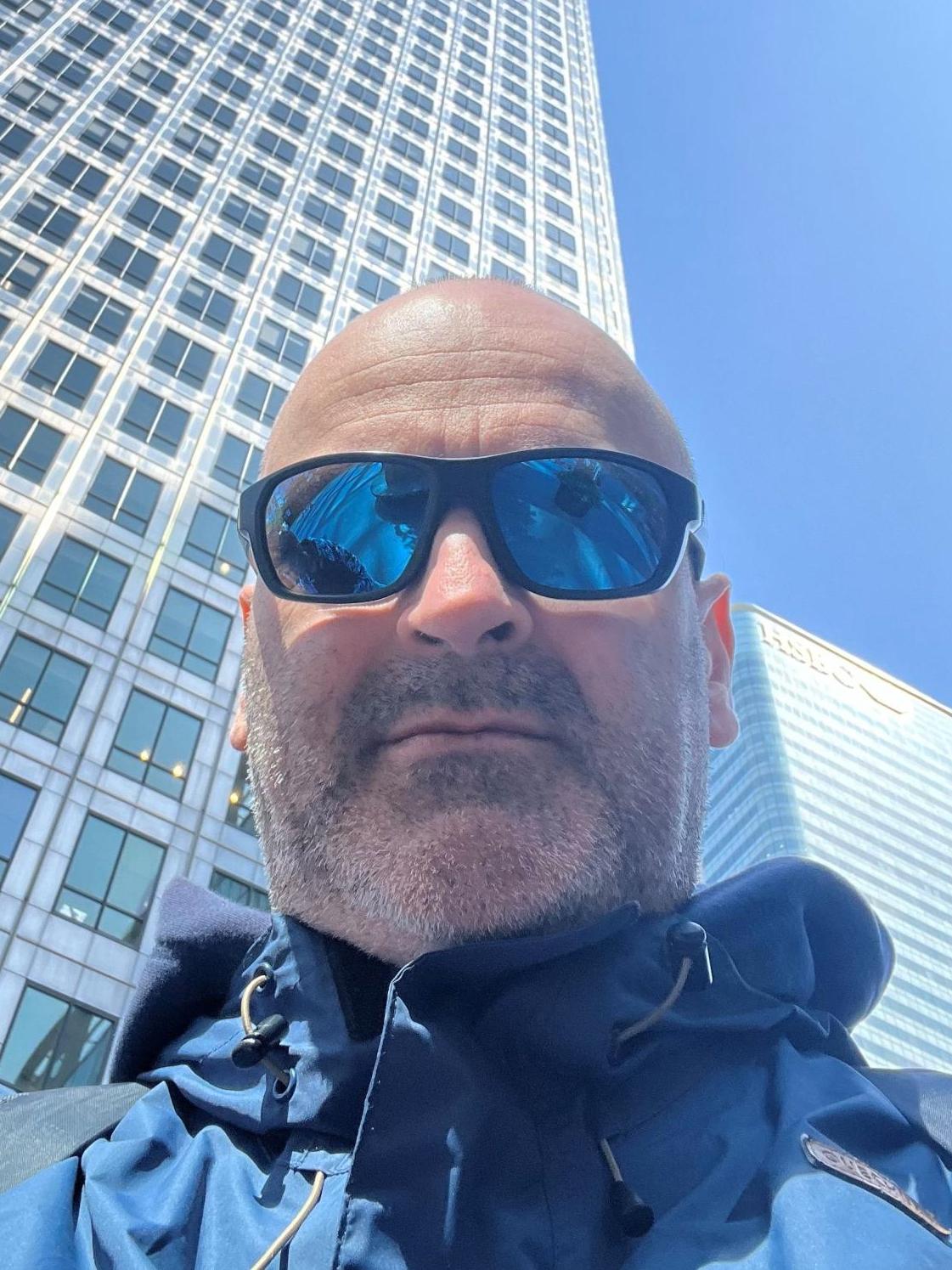 Saksofonista Marek Podkowa na tle wysokiego, szarego drapacza chmu, ubramy jest w niebieską kurtkę, nosi przeciwsłoneczne okulary, posiada lekki zarost i łysiejącą głowę